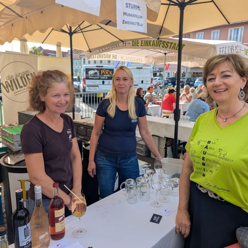 Three women stand behind a table with wine glasses and bottles, smiling at the camera. They are at an outdoor market with umbrellas and a van in the background.