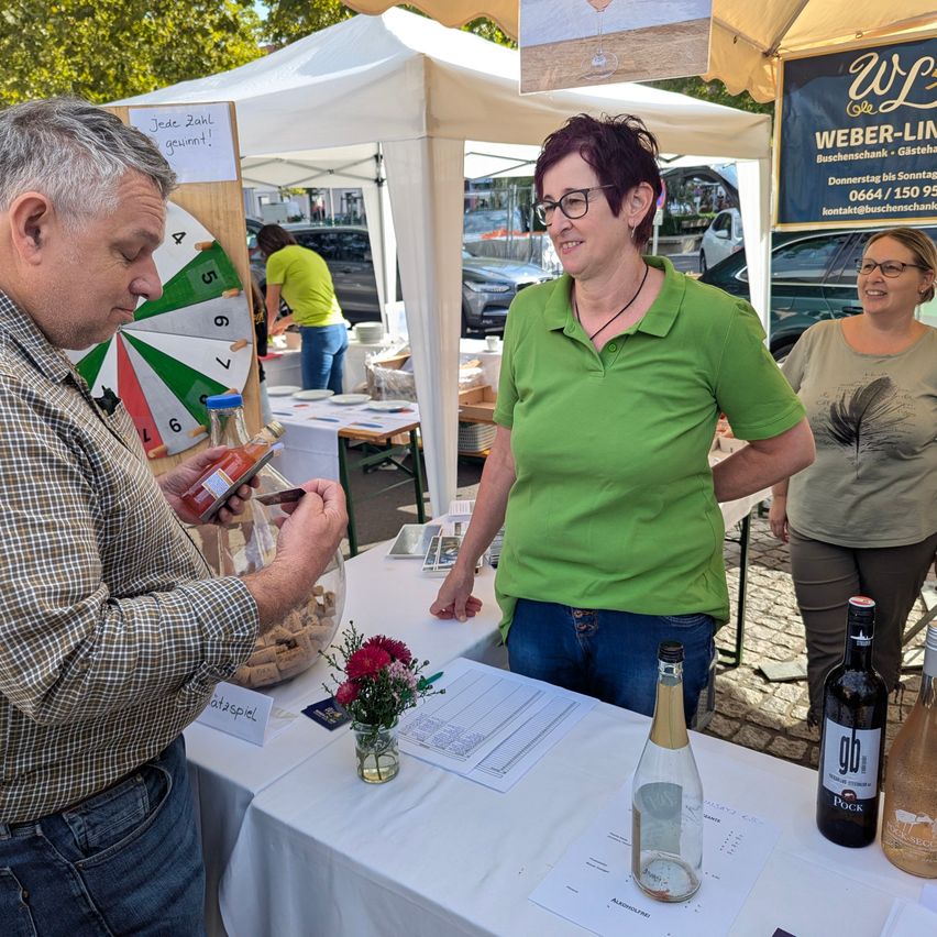 A man pours a drink into a glass while a woman in green watches. They are at a market stall with bottles and a sign.