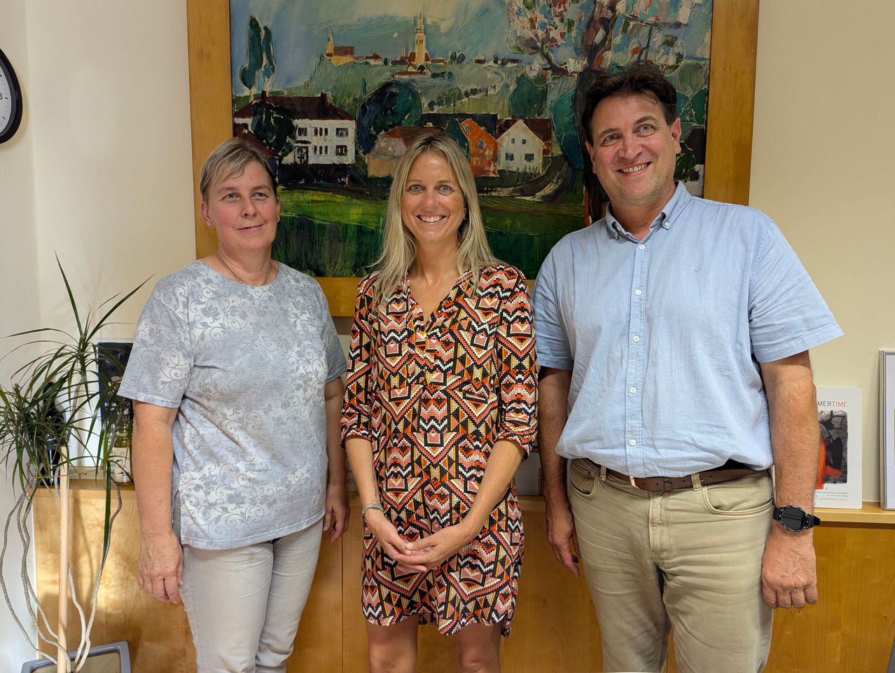 Three individuals stand together in front of a painting, smiling for the camera. The woman on the left wears a gray top, the middle woman an orange dress, and the man on the right a blue shirt.