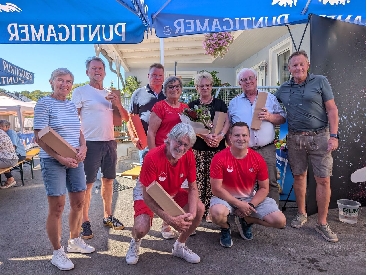 A group of people, some holding flowers and boxes, are posing for a photo under a blue canopy. The man in red is kneeling while others stand behind him.