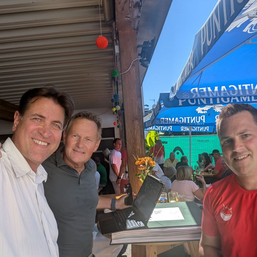 Three men are smiling and posing for a photo at an outdoor event. The man on the left wears a white shirt, the middle man wears a gray polo, and the right man wears a red shirt. Behind them, a table with a laptop and flowers is visible, and people are seated at tables under blue umbrellas.