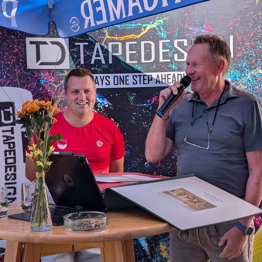 Two men stand by a table with a laptop and flowers. One man holds a microphone and smiles, while the other looks on. Behind them, a colorful banner reads 'Tapedes'.