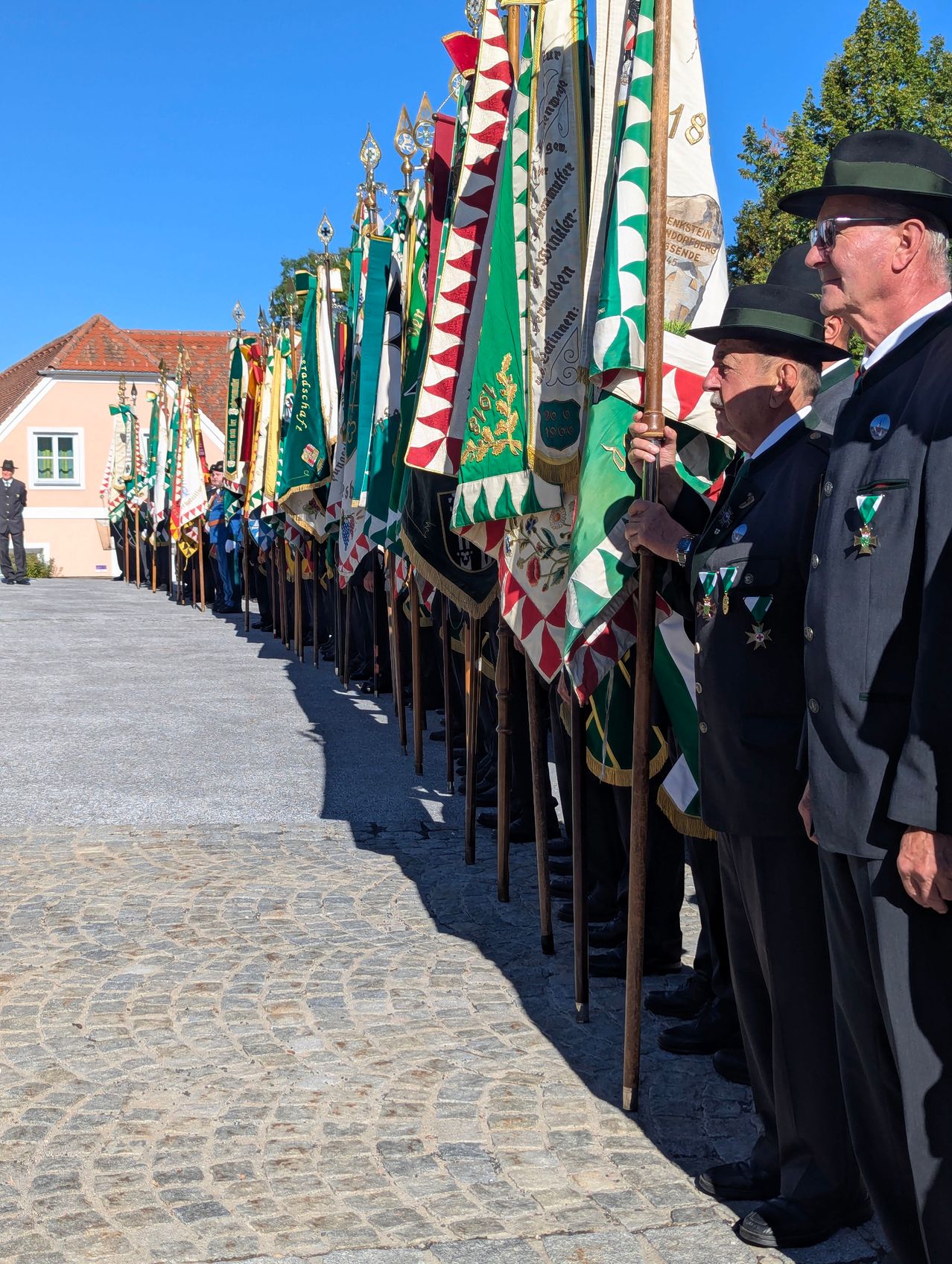 A line of uniformed individuals holding flags stand in formation, with two men in the foreground wearing hats and medals.