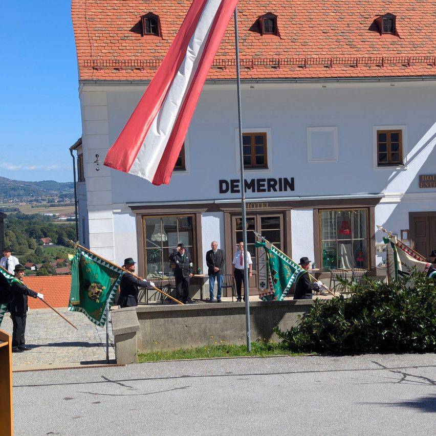 A group of people in formal attire are standing in front of a building with a flagpole. The building has the text 'DE MERIN' on it. There are green flags being held by some of the people.
