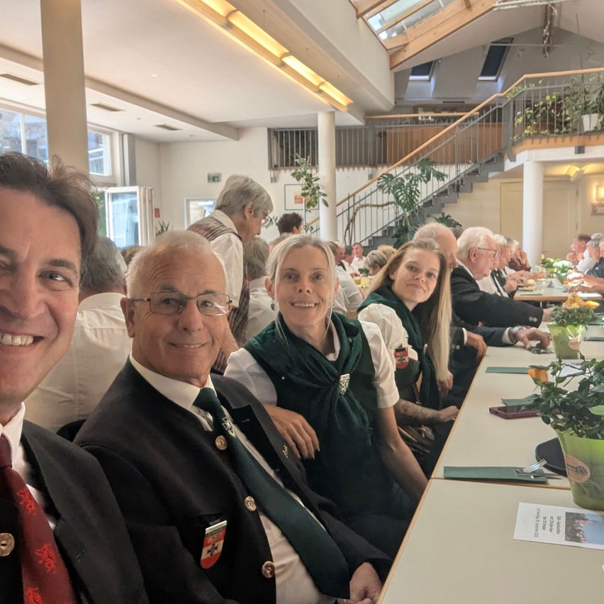 A group of people are sitting at a long table in a dining hall, smiling and posing for a photo. The table is decorated with flowers, and the room has a staircase and plants.