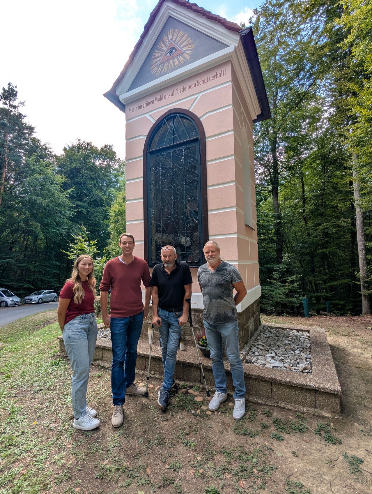 Four individuals stand in front of a small chapel in a forest, with one man using a cane.