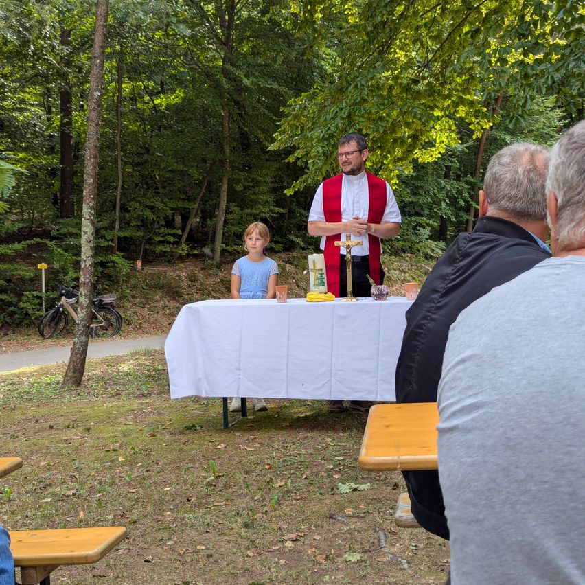 A priest stands at a table in a forest, surrounded by a small group of people. A young girl stands next to him.