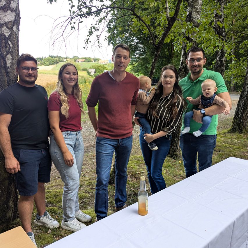 A group of people, including adults and children, are standing in a park, smiling and posing for a photo. A table with a bottle is in front of them.