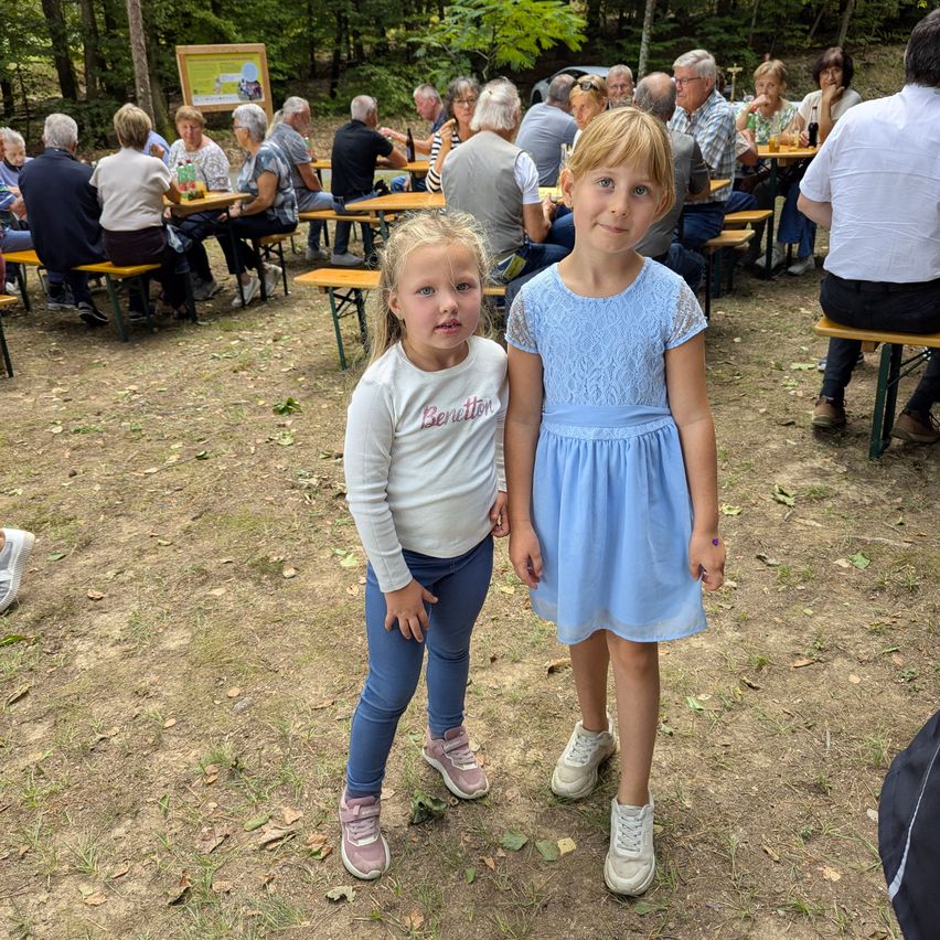 Two young girls stand in a park with an outdoor dining area in the background. One girl wears a white shirt with the word 'Benetti' and the other wears a blue dress.