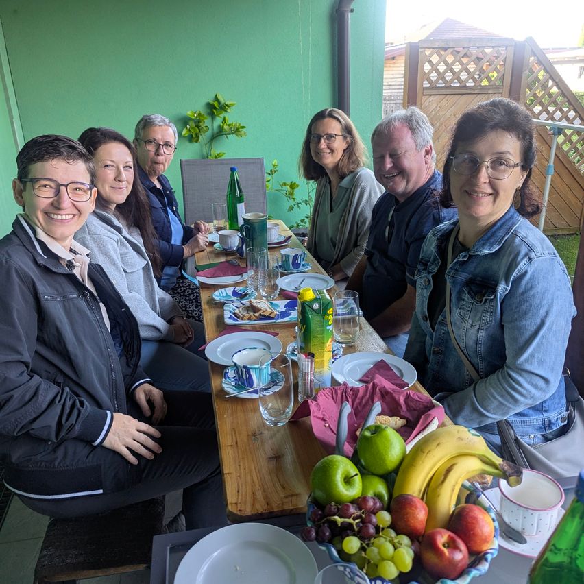 A group of people are sitting at a table with food, drinks, and fruit. They appear to be having a meal together in a garden.