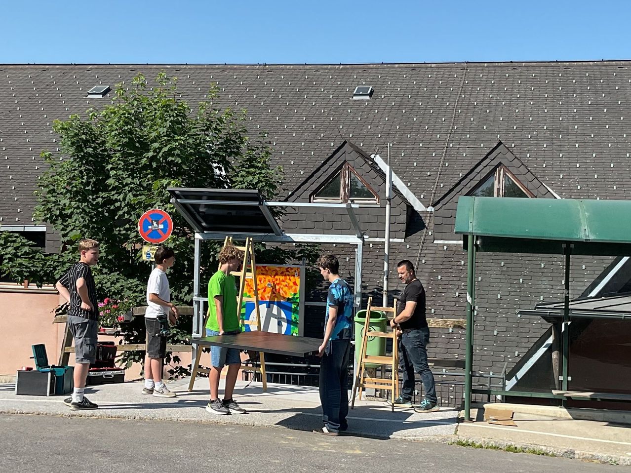 Five men are setting up a canvas on a stand in front of a building. A green trash bin and a sign are nearby.