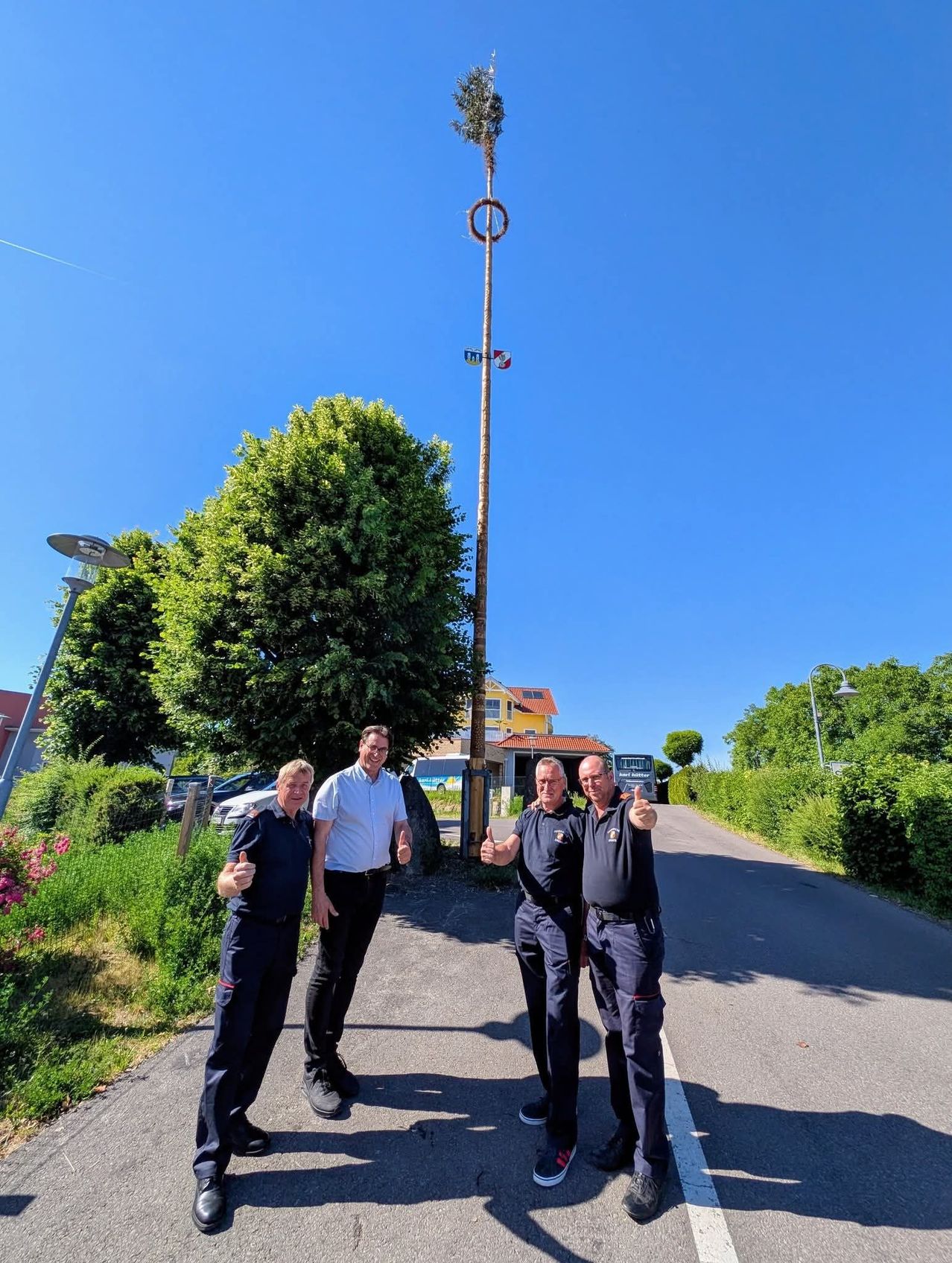 Three men stand on a road with a pole, trees, and parked cars in the background. They are smiling and giving thumbs up.