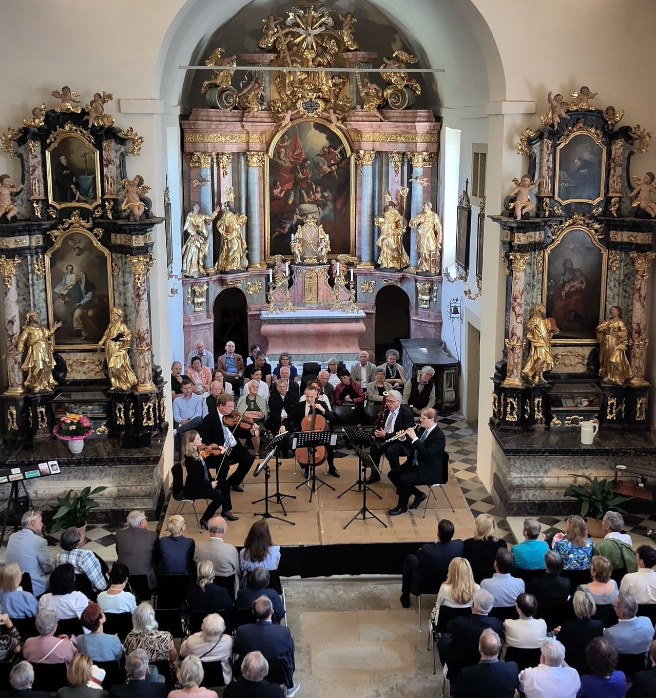 A classical music ensemble performs in a church with a large audience. Musicians play violins and flutes on stage, with ornate altars and paintings in the background.