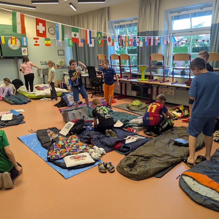 Kinder sitzen und stehen um ein Klassenzimmer herum, einige mit Schlafsäcken und andere mit Gegenständen in den Händen. Eine Frau steht im Hintergrund.