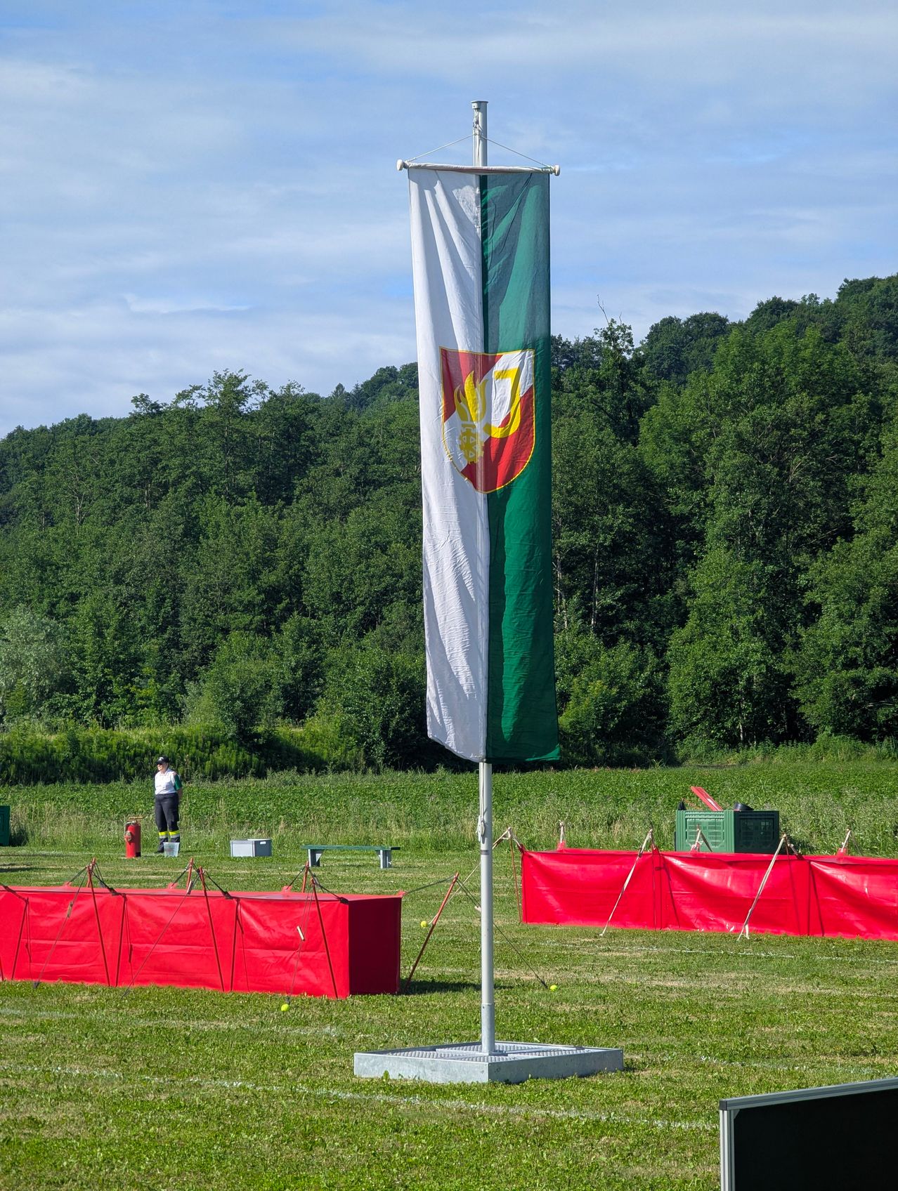 Ein großes Banner mit Logo weht auf einem Pfahl in einem Grasfeld mit roten Barrieren. Eine Person steht in der Nähe eines Feuerlöschers. Bäume und Büsche im Hintergrund.