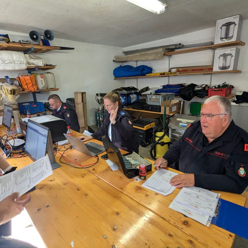 Eine Gruppe von Menschen in Uniformen sitzt um einen Tisch mit Laptops und Papieren. Sie befinden sich in einem Raum mit Regalen und Aufbewahrungsboxen. Sie scheinen etwas zu besprechen.