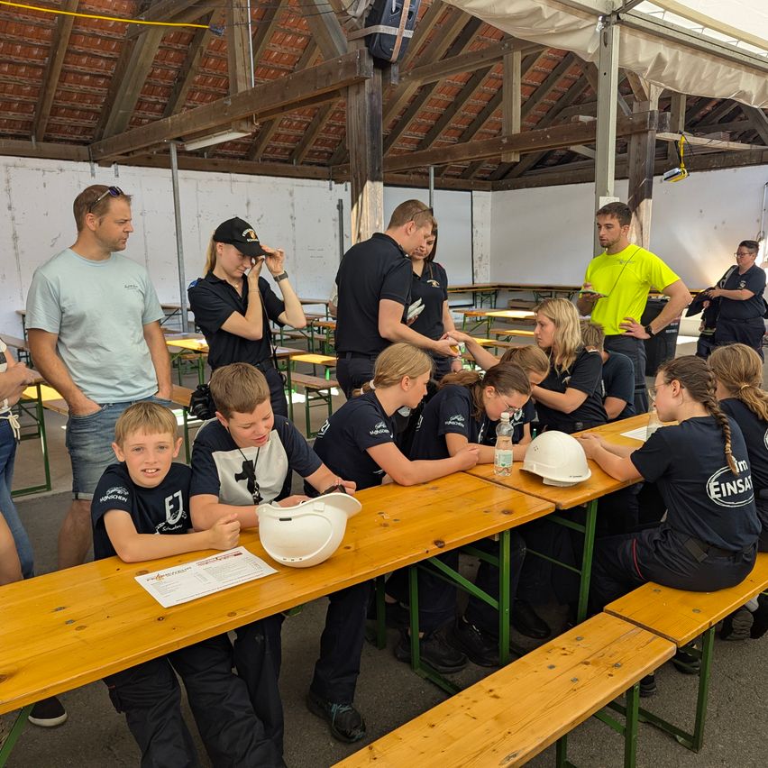 Mehrere Personen, darunter Kinder, sitzen an einem langen Holztisch in einem Pavillon, möglicherweise an einer Veranstaltung teilnehmend. Sie tragen alle Uniformen und einige haben Helme auf dem Tisch. Erwachsene stehen um sie herum.