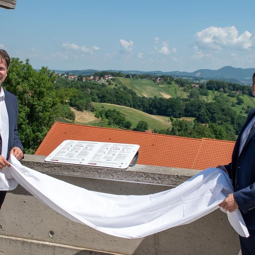 Zwei Männer in Anzügen stehen auf einem Balkon, halten ein weißes Tuch und lächeln. Dahinter ist eine Speisekarte auf einer Betonfläche mit Blick auf Hügel und ein Dorf platziert.