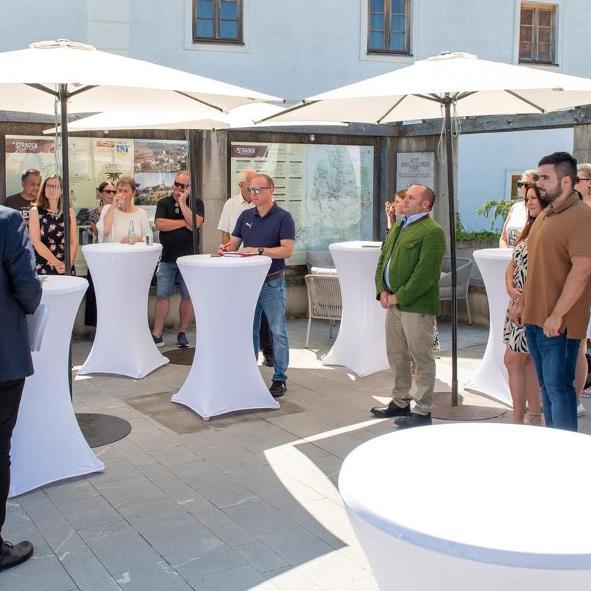 Eine Gruppe von Menschen steht in einem Außenbereich, einige sitzen auf Stühlen unter Schirmen, und ein Mann steht und spricht mit einem anderen Mann.