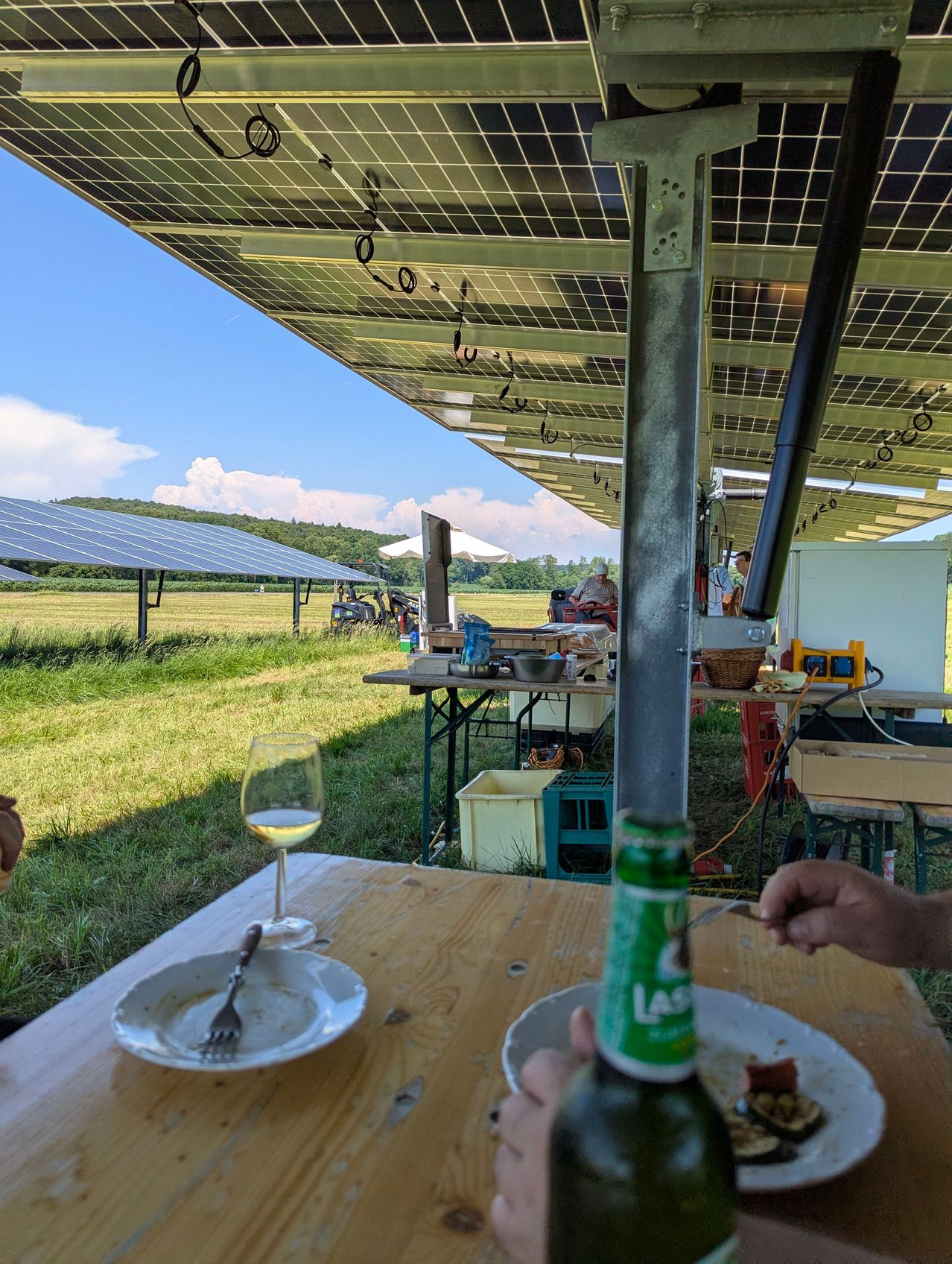 Eine Outdoor-Installation mit Solarpanelen auf einem Feld. Ein Tisch hat ein Glas, eine Gabel und einen Teller mit Essen. Ein weiterer Tisch ist mit verschiedenen Gegenständen besetzt. Eine Person öffnet eine Flasche.