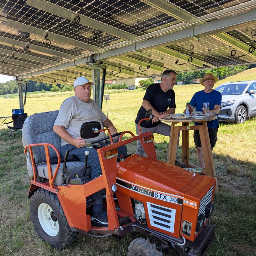 Drei Männer sitzen unter einem solarbetriebenen Vordach, einer fährt einen roten Fiat-Traktor, ein anderer hat ein Glas Wein und der dritte hält einen Becher.
