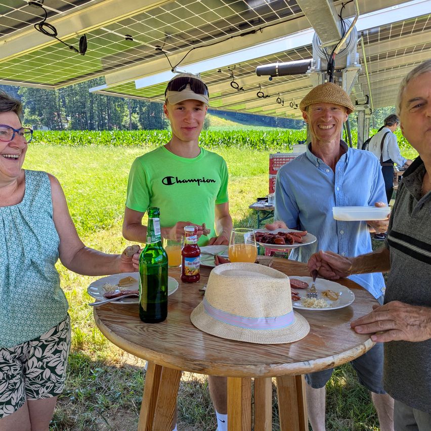Vier Personen stehen um einen Tisch mit Essen und Getränken. Eine Person hält einen Teller mit Essen, eine andere hat eine Flasche. Sie sind draußen in der Nähe von Solarmodulen.