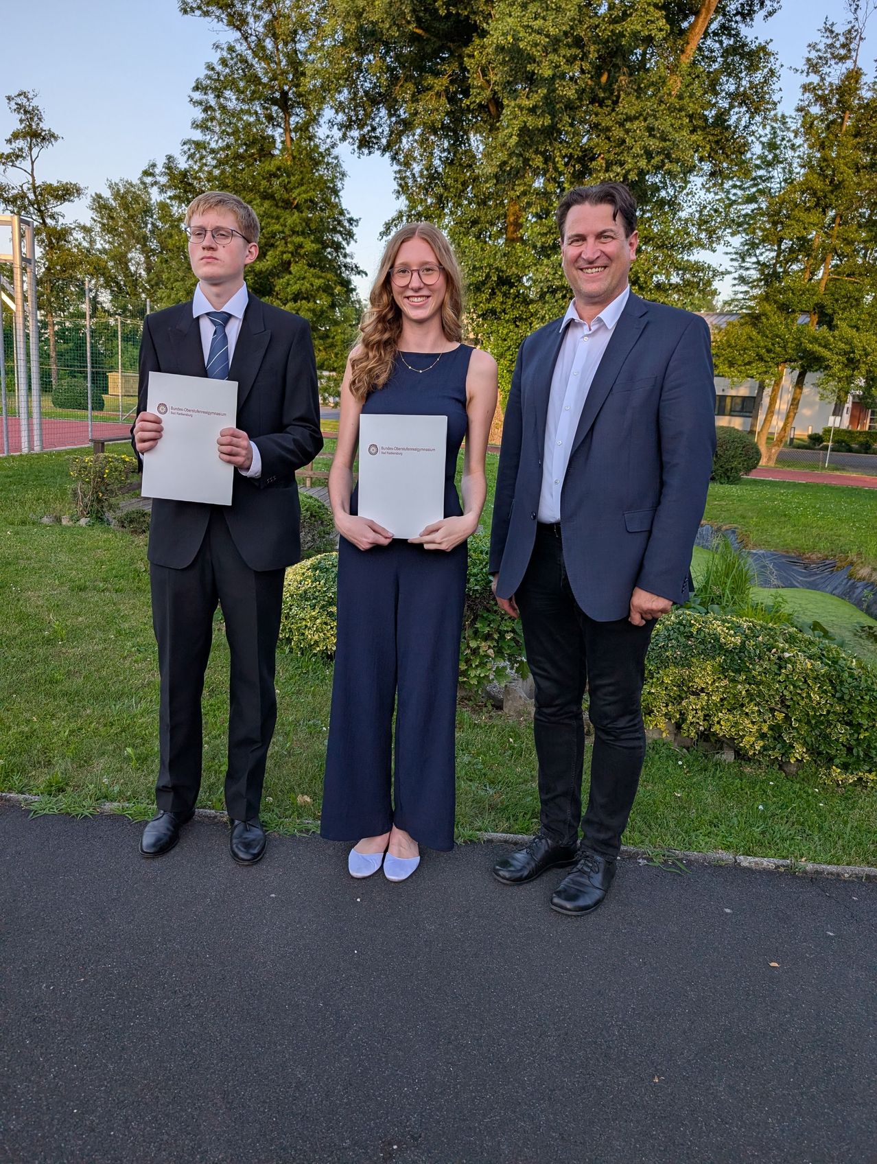Drei Personen stehen in Anzügen draußen und halten Papiere in der Hand. Ein Mann links, eine Frau in der Mitte und ein Mann rechts. Sie stehen auf einer gepflasterten Straße in einem Park.