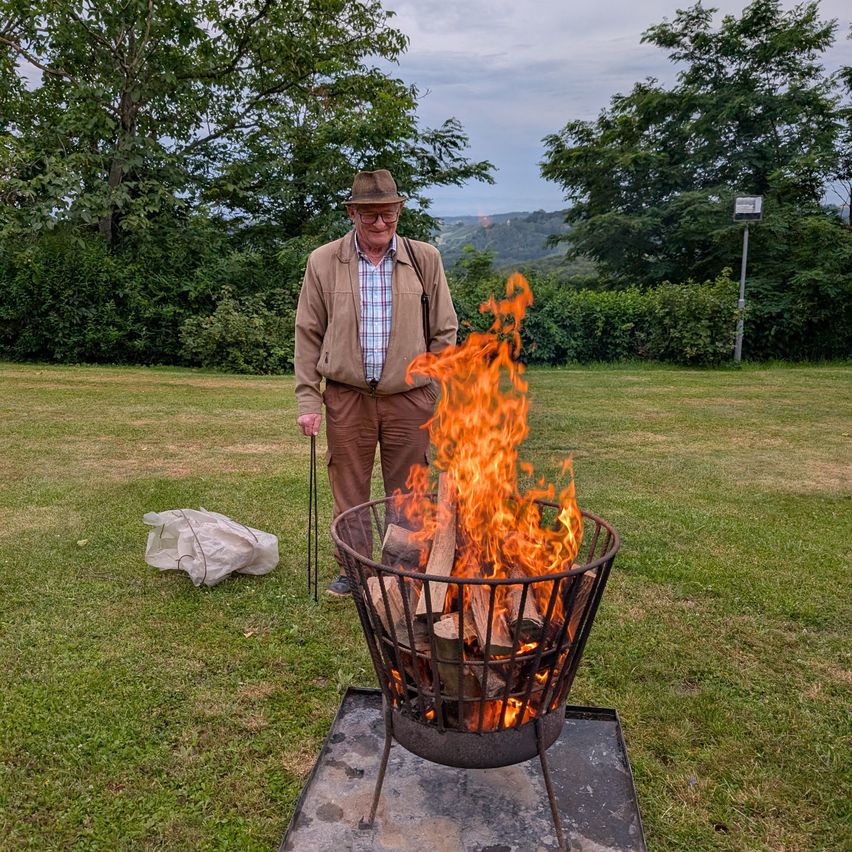Ein älterer Mann in einer braunen Jacke steht neben einem brennenden Feuerkorb in einem Grasfeld mit freier Sicht auf die Berge.
