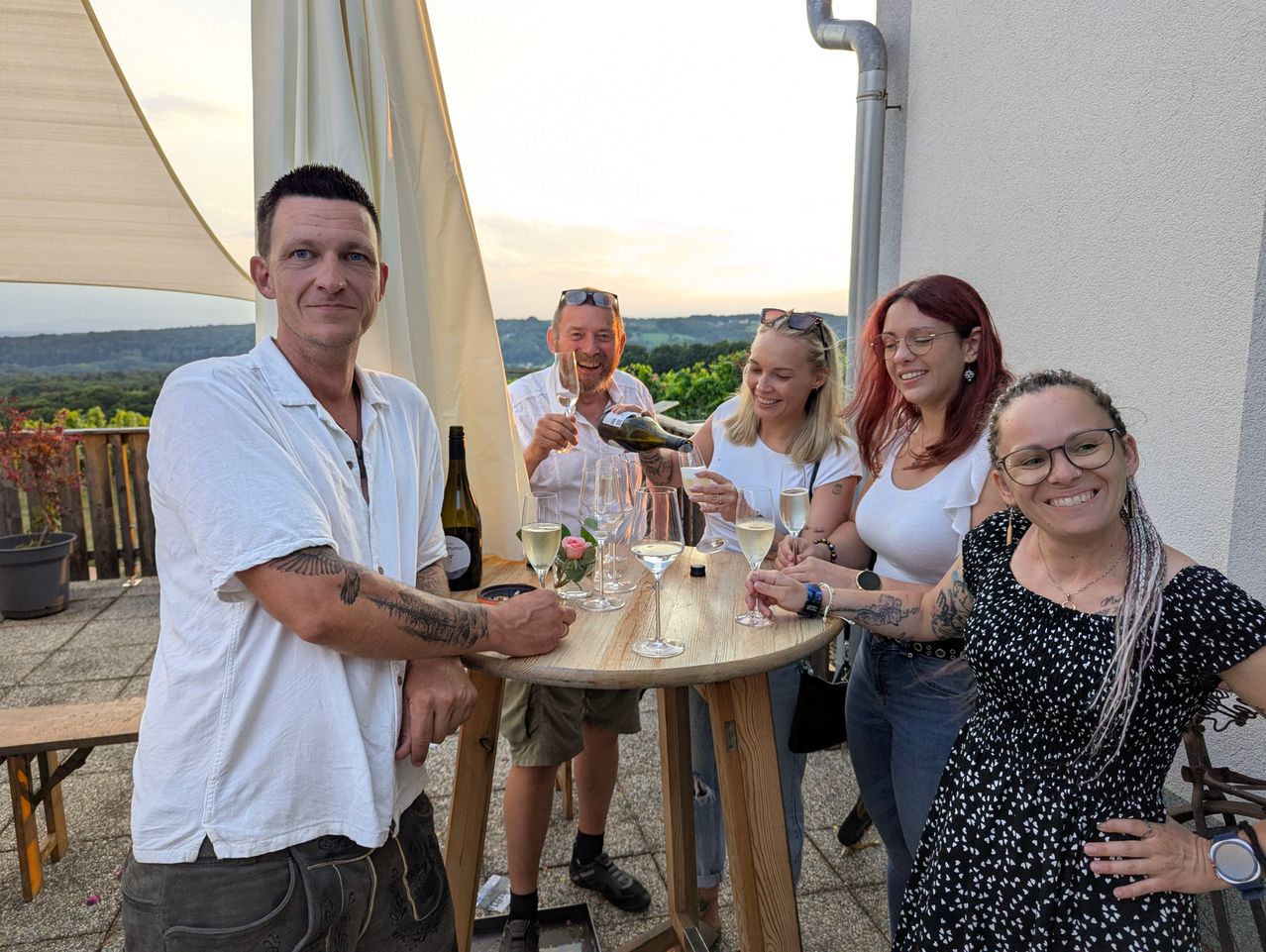 Eine Gruppe von Menschen steht um einen Tisch herum, lächelt und hält Weingläser. Ein Mann schenkt Wein in die Gläser. Sie stehen auf einer Terrasse mit Blick auf den Sonnenuntergang.