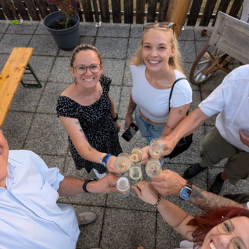 Eine Gruppe von vier Personen stößt mit Champagnergläsern in einem Kreis an, lächelt und schaut zur Kamera. Der Hintergrund scheint draußen zu sein mit einem Holzzaun und einer Holzbank.