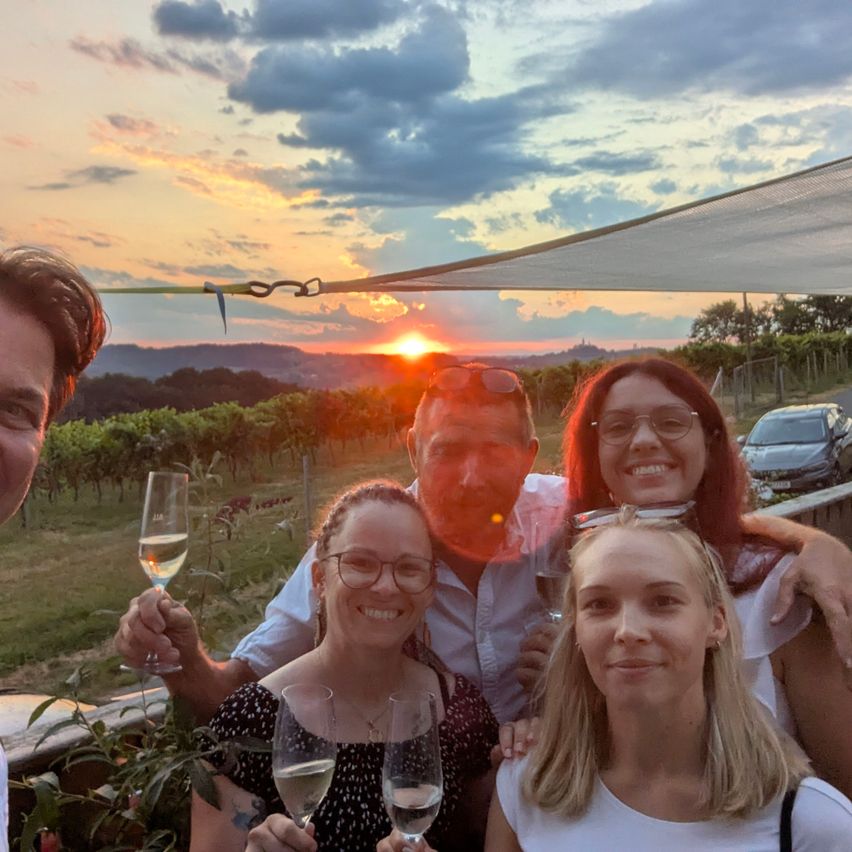 Fünf Menschen lächeln und posieren für ein Foto in einem Weinberg mit einem Sonnenuntergang im Hintergrund. Sie halten Weingläser und scheinen zu feiern.