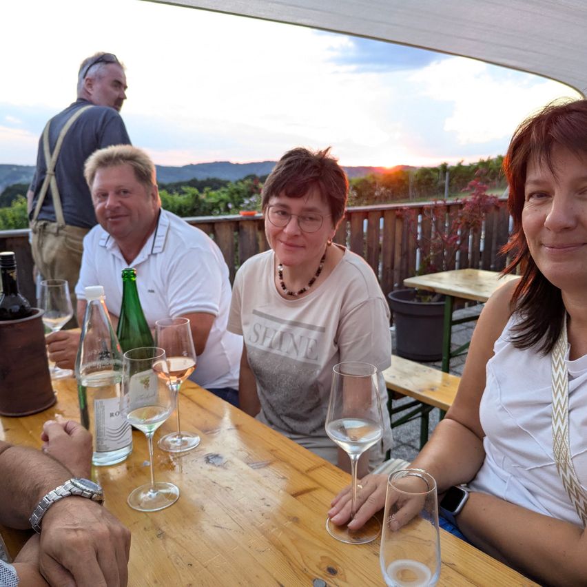 Vier Personen sitzen an einem Tisch und lächeln für ein Foto. Der Tisch hat Weingläser, Flaschen und einen Topf. Dahinter ist ein Holzzaun und eine Sonnenuntergangskulisse zu sehen.
