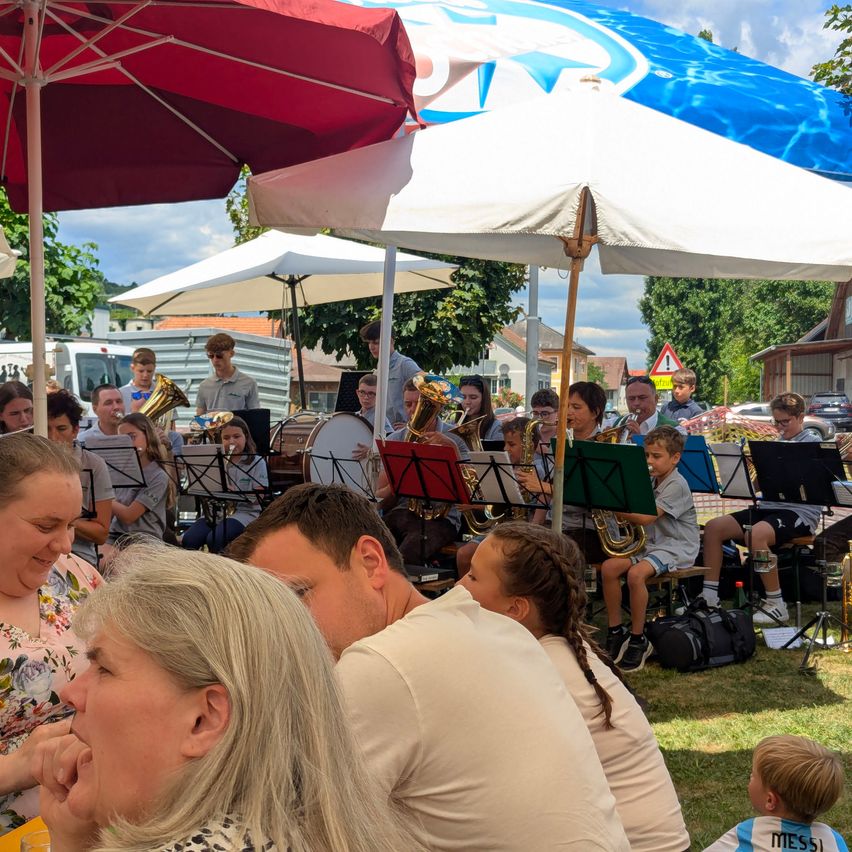 Eine Schulband spielt auf einem Rasenfeld, während mehrere Menschen zuschauen. Die Band besteht aus Blech- und Perkussionsinstrumenten. Schirme spenden den Zuschauern Schatten.