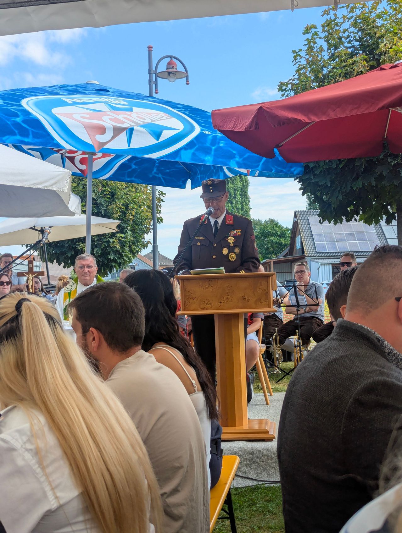 Ein Militäroffizier in Uniform steht unter einem Schirm an einem Podium und spricht zu einer Menschenmenge. Ein Mann spielt im Hintergrund Musik.