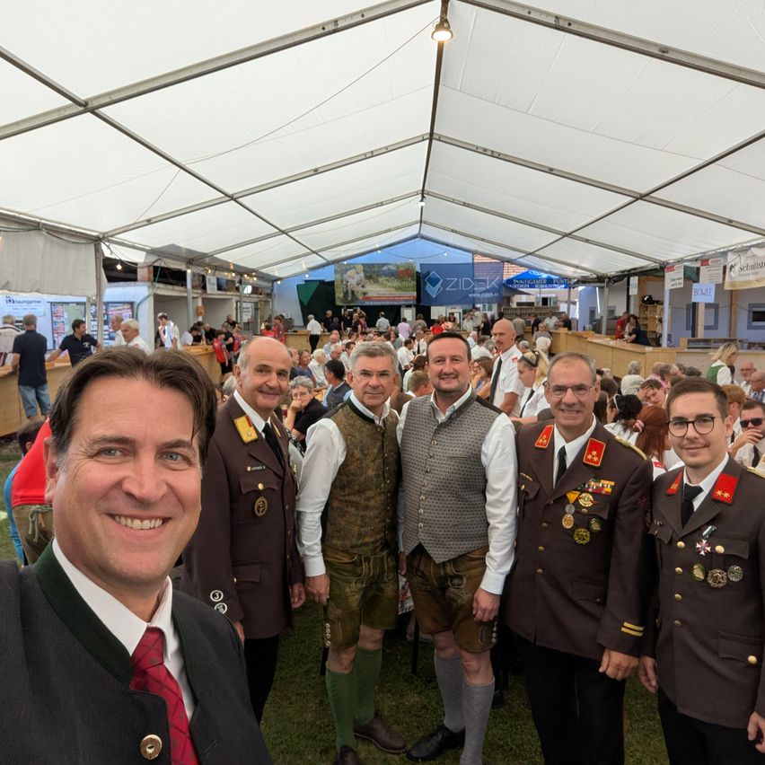 Eine Gruppe von Männern in traditioneller deutscher Kleidung, die lächelnd für ein Foto bei einem Festival posieren. Dahinter versammelt sich eine Menschenmenge in einem Zelt.