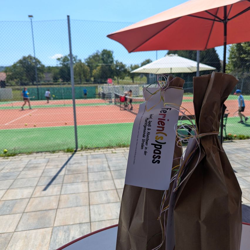 Ein brauner Papierbeutel mit einem weißen Etikett, das Ferienpass liest, steht auf einem Tisch unter einem roten Schirm. Im Hintergrund ist ein Tennisplatz mit Spielern und einem weißen Schirm zu sehen.