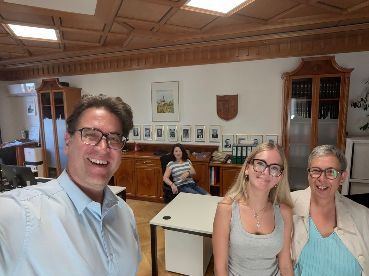 Drei Menschen lächeln in einem Büro. Ein Mann in einem blauen Hemd steht vorne, während zwei Frauen hinter ihm stehen. Hinter ihnen sitzt eine Frau an einem Schreibtisch. Der Raum hat Holzschränke, ein Bilderrahmen und ein Schild an der Wand.