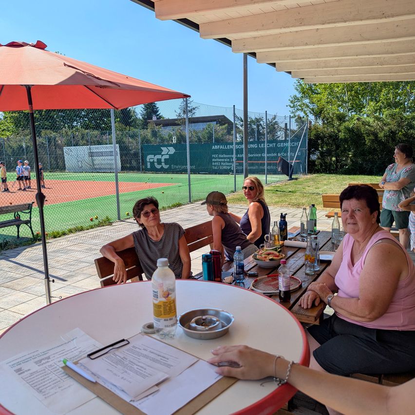 Eine Gruppe von Frauen sitzt an einem Tisch in einem Café im Freien und genießt Essen und Getränke. Hinter ihnen ist ein Tennisplatz mit einem Zaun und Bäumen zu sehen.
