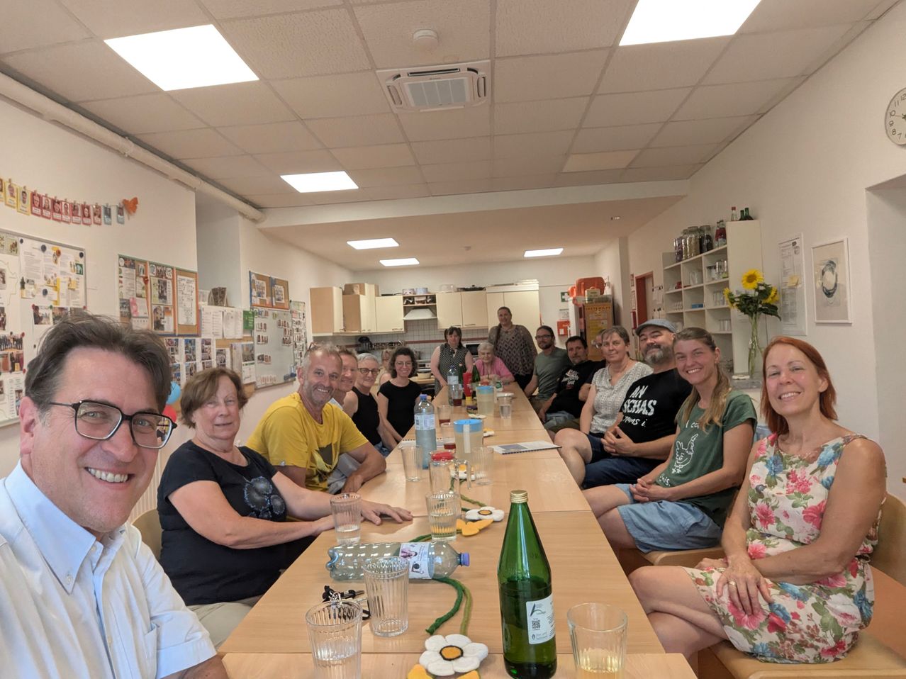 Eine Gruppe von Menschen sitzt um einen Tisch in einem Raum mit weißer Decke und Wänden. Auf dem Tisch stehen Flaschen, Gläser und eine Blumenvase. Einige Leute tragen Brillen und Hüte.