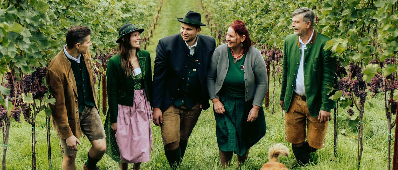 Fünf Personen stehen in einem Weinberg, gekleidet in traditioneller Kleidung. Die beiden Männer tragen grüne Jacken und Hüte, während die Frauen Kleider tragen. Ein Hund läuft in der Nähe. Hinter ihnen sind ein Haus und eine Scheune zu sehen.