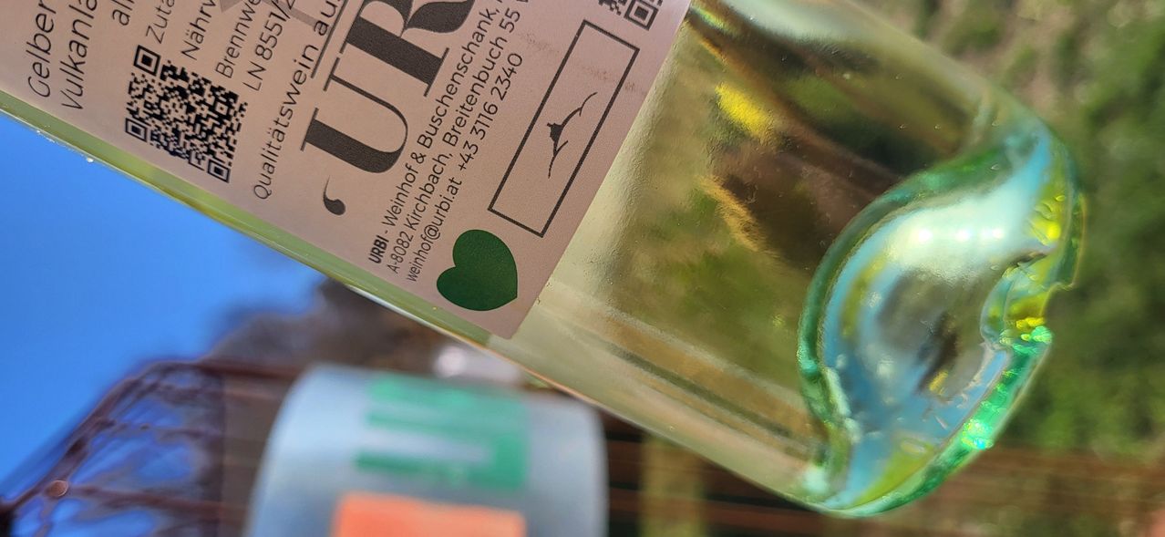 Eine Nahaufnahme einer Weinflasche mit einem grünen Herz-Aufkleber auf dem Etikett. Das Etikett trägt den Namen 'URSI' und einen QR-Code. Die Flasche hat einen transparenten Körper mit einem grünlichen Farbton.
