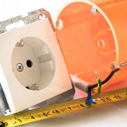 Eine Steckdose liegt auf einem Messband, mit angeschlossenen Drähten und einer offenen Anschlussdose in der Nähe.