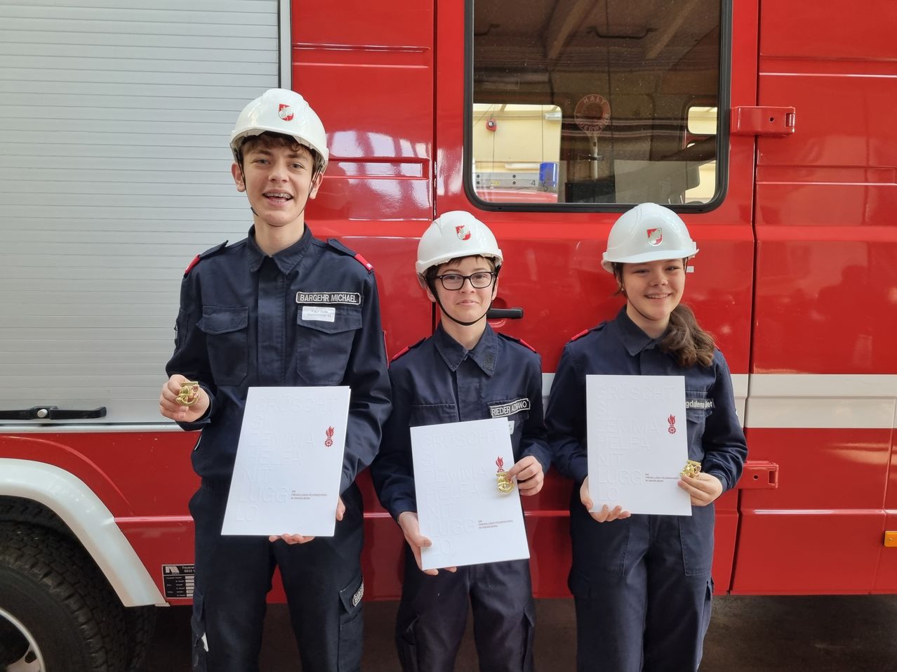 Drei junge Personen in Marineblau-Uniformen und weißen Helmen stehen vor einem Feuerwehrwagen, jeder hält ein Zertifikat und eine Medaille.