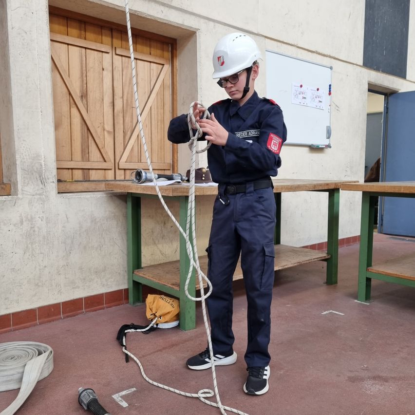 Eine Person in einer blauen Uniform hält ein Seil in einem Raum mit einer Tafel, Tischen und einer Tür.