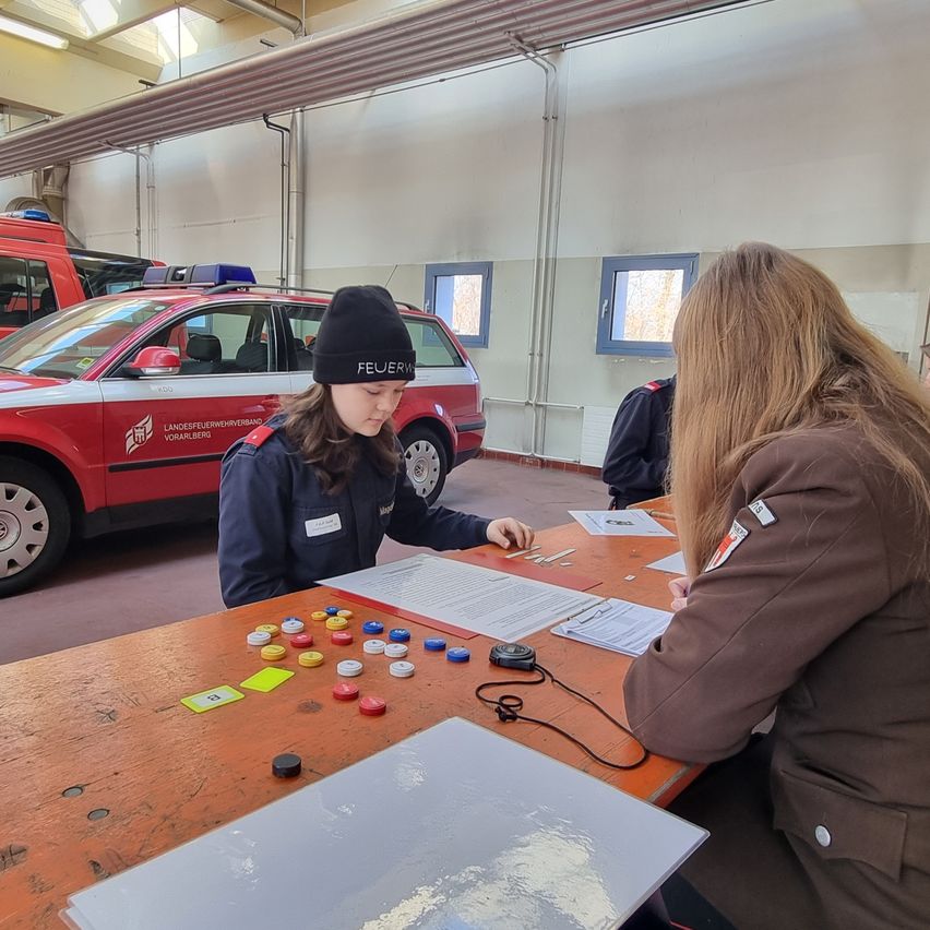 Zwei Frauen in Uniformen sitzen an einem Tisch in einer Garage. Ein Feuerwehrauto ist hinter ihnen geparkt.