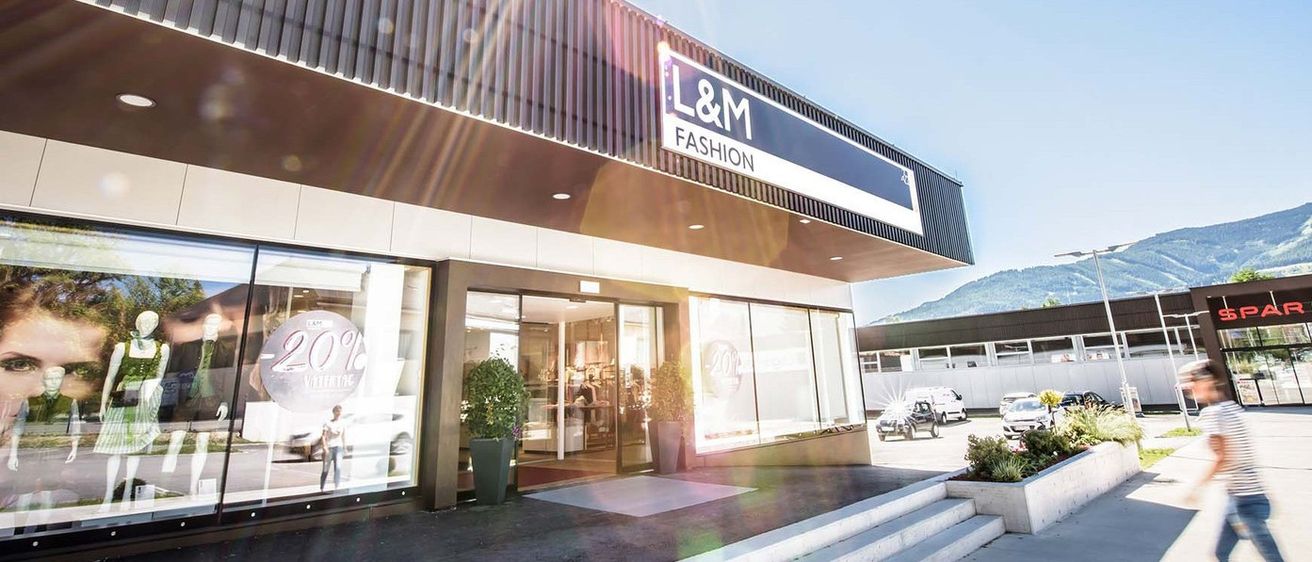 Der Eingang des L&M Fashion-Geschäfts ist mit einem Schild oben zu sehen. Autos sind davor geparkt. Topfpflanzen stehen an den Seiten.