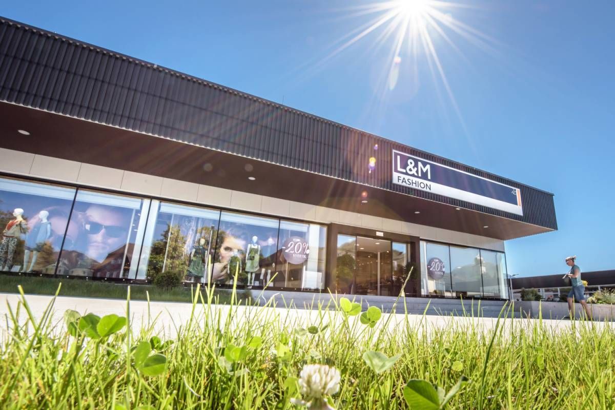 Ein Gebäude mit 'L&M FASHION' Schild, reflektiert Sonnenlicht. Fenster zeigen Puppen und Kleidung. Ein 20% Rabattschild ist sichtbar. Heller Himmel oben.