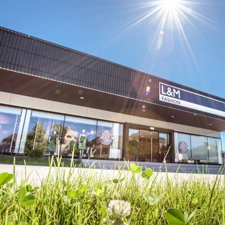 Ein Gebäude mit 'L&M FASHION' Schild, reflektiert Sonnenlicht. Fenster zeigen Puppen und Kleidung. Ein 20% Rabattschild ist sichtbar. Heller Himmel oben.