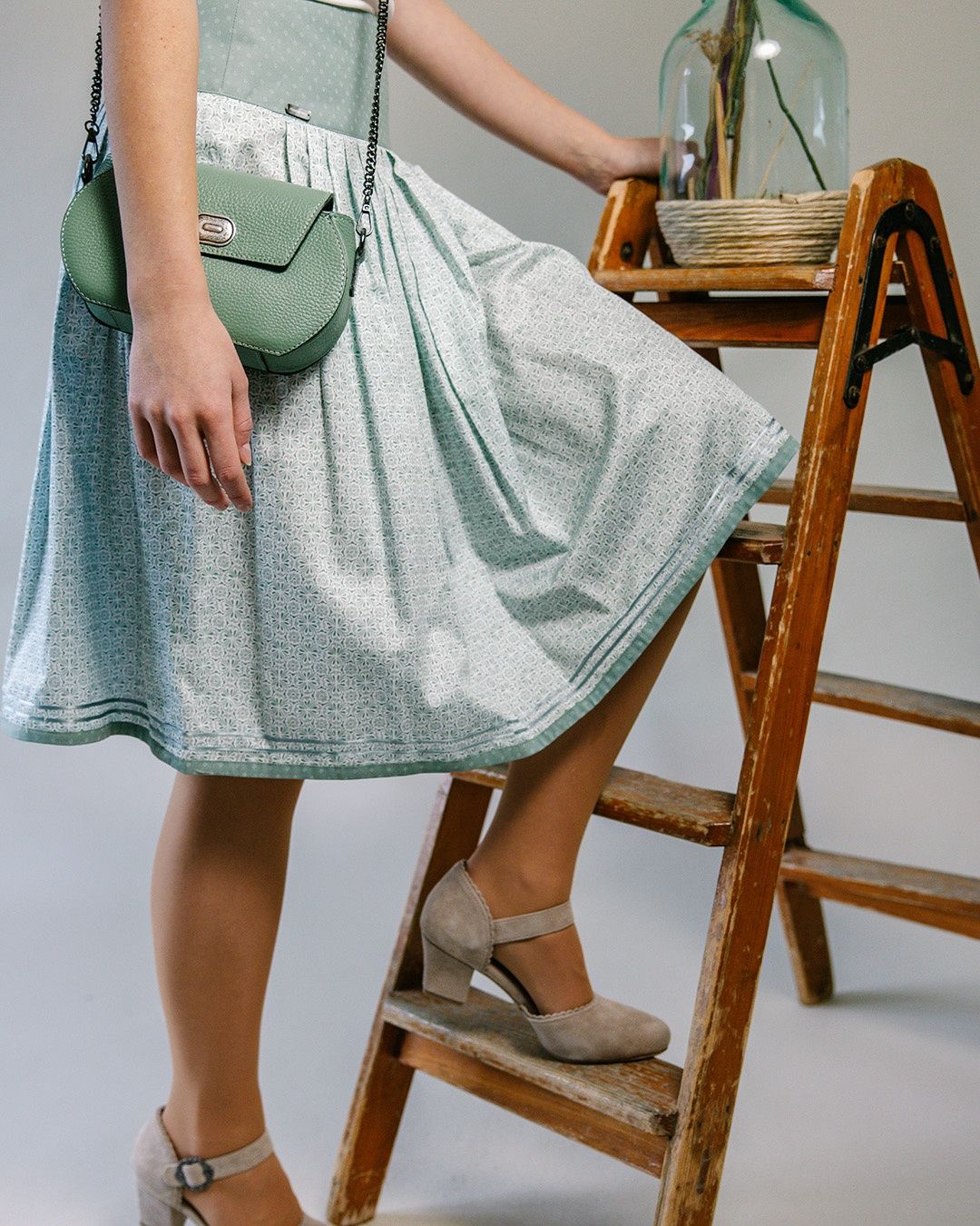 Eine Frau steht auf einer Holzleiter, trägt einen Rock und eine grüne Handtasche. Der Rock ist grün und weiß mit Muster. Sie trägt beigefarbene High-Heels.