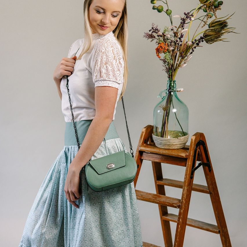 Eine Frau in einem weißen Oberteil und einer blauen Rock posiert mit einer grünen Umhängetasche. Hinter ihr steht eine hölzerne Leiter mit einer Glasvase mit getrockneten Blumen.